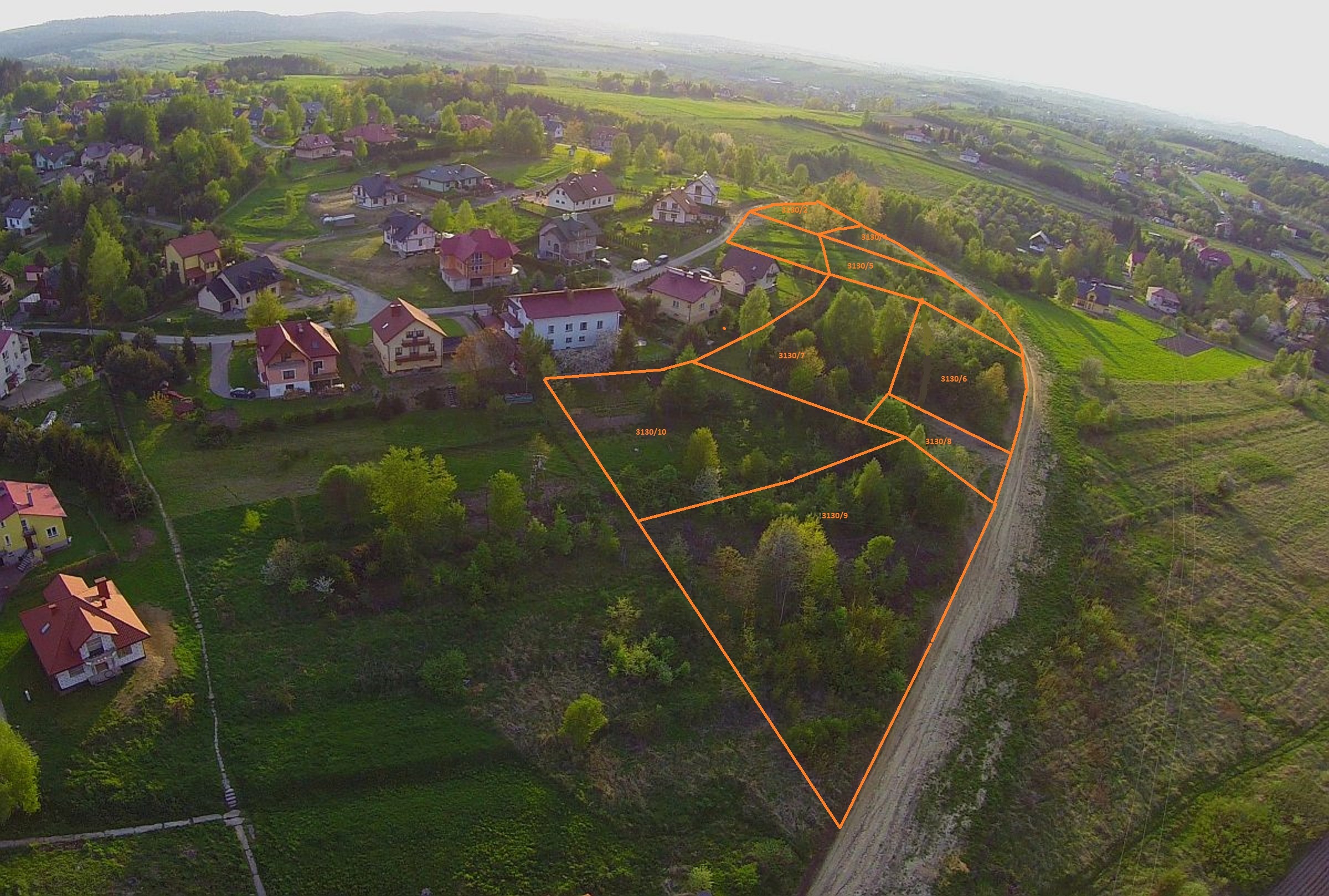 Zdjęcie lotnicze terenu wiejskiego z domami i działkami, oznaczonymi pomarańczowymi liniami i numerami. Widok na zielone wzgórza.